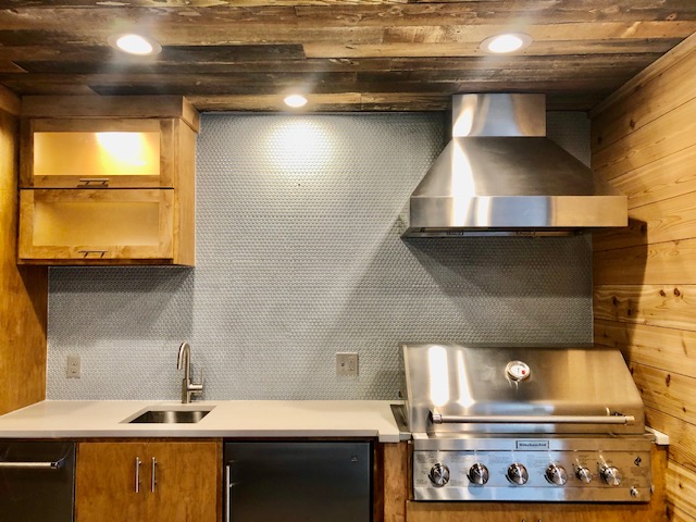 New outdoor kitchen with penny tile backsplash, quartz countertop, custom made cabinets, LED lighting, vent, and gas grill.