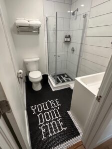 Renovated bathroom with black tile flooring, glass shower, and a custom 'You're Doing Fine' design in a 1927 Norman house.