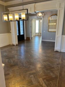 Interior view of a room showcasing new wood floors with brass inlay and a combination of traditional and herringbone patterns. The space features white molding, modern lighting, and large windows.